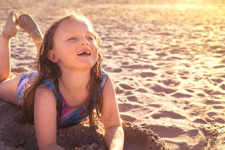 Happy little girl in a swimsuit lies on the beach and smilesの写真素材