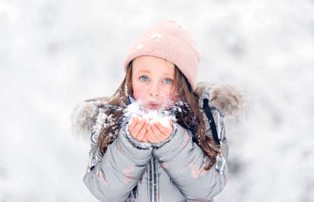 A beautiful blue-eyed girl blows on the snow in her palms in winter, a portrait of a childの写真素材