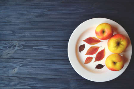Apples in a white plate on a wooden table. Ripened apples. Autumn leaves decor.