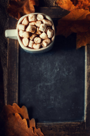 Autumn, fall leaves on vintage Empty blank black chalkboard with hot cup of coffee, marshmallows  and  copyspace. Cozy Autumn Still Life Background. Autumn red and orange leavesの写真素材