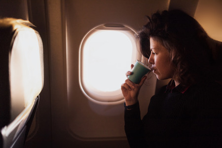 Young Caucasian  woman drinking coffee  inside airplane sitting on passenger seat near window. Happy Female airplane passanger enjoying her flightの写真素材