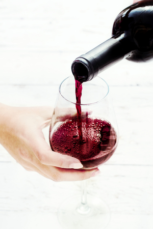 Woman hand Pouring red  wine from bottle into the wine glass on white wooden background with copyspaceの写真素材
