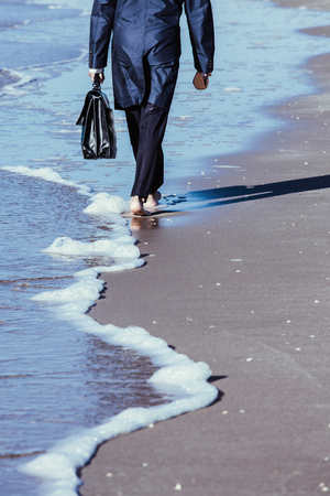 A lone man in a raincoat, with a briefcase strolling along the edge of the water. Businessman walking bare foot on sandy beach near the sea, after work.の写真素材