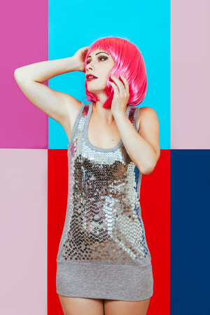 Beautiful girl with pink hair dressed in silver dress on a multicolored graphic background. A happy woman emotionally touches head her hands.の写真素材
