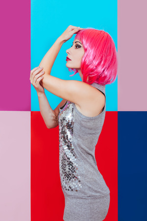 Beautiful girl with pink hair dressed in silver dress on a multicolored graphic background. A happy woman emotionally touches head her hands.の写真素材
