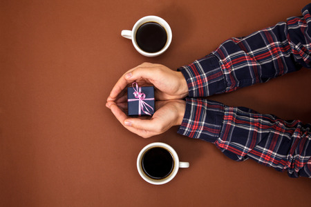 The male hands are closed on a brown background. The guy gives a gift to a girl. Men's hands next to two cups of coffee and with gift.の写真素材
