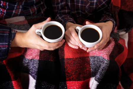 Young couple's hands with two cups of hot coffee. Man and woman together in a warm blanket.の写真素材