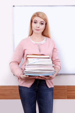 Portrait of blonde female teacher in classroom against the white school board. A young surprised teacher holds a lot of books in her hands.の写真素材