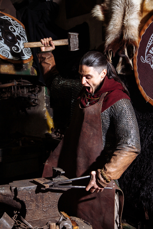 Viking forges weapons and swords in the smithy. A man in a warrior's clothes is in the smithy on the background of fire.の写真素材