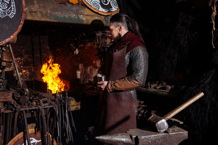 Viking forges weapons and swords in the smithy. A man in a warrior's clothes is in the smithy on the background of fire.の写真素材