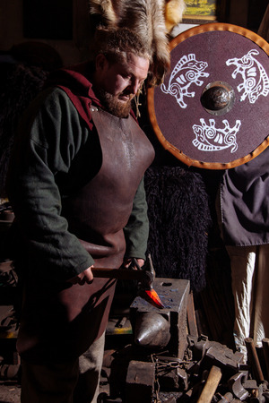 Viking forges weapons and swords in the smithy. A man in a warrior's clothes is in the smithy on the background of fire.の写真素材