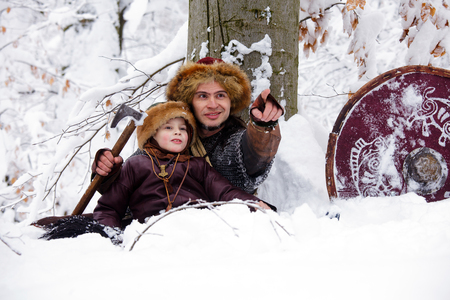 Father and son Vikings in a deep winter forest and the father teaches his son to be a real Viking. Viking warrior with chain mail leather spear walking in winter woods before battle, scandinavian traditional clothing for lumberjack, viking concept.の写真素材