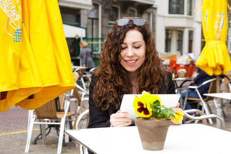 Young girl with long curly hair sitting in a street cafe with coffee and juice in the spring. A beautiful woman uses a tablet to communicate. Street life and travel in the old town.の写真素材