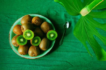 A lot of kiwi on the green table. Healthy fruit kiwi on the plate with tropical leaves monstera. Tropical fruit kiwi. Still life.の写真素材