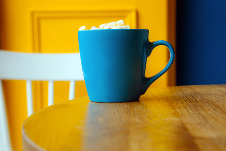 Blue cup of coffee on yellow background. A cup of chocolate sprinkled on top with a little white and a pink marshmallow.の写真素材