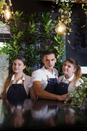 A cute young waiters in a white T-shirts and a blue aprons holding a cup of coffee in hands. Portrait of confident young male and female barista.の写真素材
