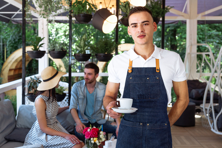 A cute young waiter in a white T-shirt and a blue apron holding a cup of coffee in his hands. Portrait of confident young male barista serving coffee to customer at cafe.の写真素材