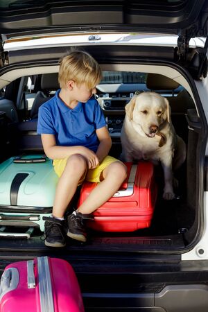 Boy are loading multicolored suitcases in the trunk of car. The kid sits in the open trunk of a car with a labrador and suitcases.の写真素材