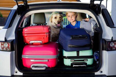 Boy are loading multicolored suitcases in the trunk of car. The kid sits in the open trunk of a car with a labrador and suitcases.の写真素材