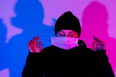 Woman in medical mask on the pink and blue background. Sick woman average age in cap on the stereet with neon lights.の写真素材