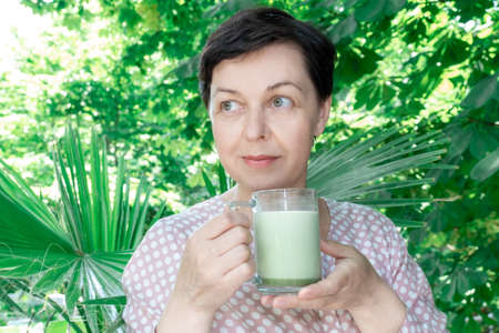 Middle Aged Woman hold glass cup of green Matcha Latte Coffee or Tea on the Tropical background. Concept of healthy food in menopause period.の写真素材