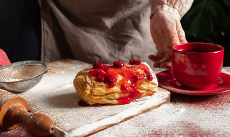Female Hands Holding Red Cup of coffee near Puff staffed with plum or red currant jam on the table.の写真素材