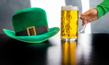 Woman's hand holding glass of beer near St. Patrick's day hat of a leprechaun on the black backgroundの写真素材