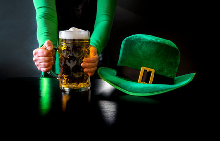 Woman's hand holding glass of beer near St. Patrick's day hat of a leprechaun on the black backgroundの写真素材