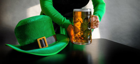 Woman's hand holding glass of beer near St. Patrick's day hat of a leprechaun on the black backgroundの写真素材