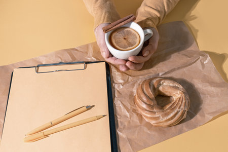Hands Holding cup of tea or Mulled Wine near Resume Sheet and Cake on the Set Sail Champagne Color Background, top view.の写真素材