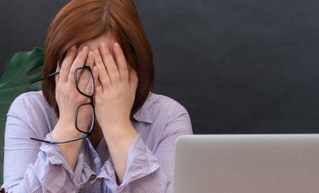 Upset Woman closed your face of hands near her workplace with laptopの写真素材
