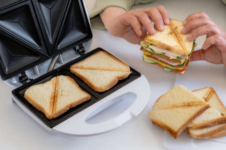 Hands hold cooked fresh sandwich with cheese, bacon and vegetable near toster on the white table.の写真素材