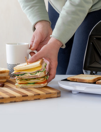Hands hold cooked fresh sandwich with cheese, bacon and vegetable near toster and cup of coffeeの写真素材