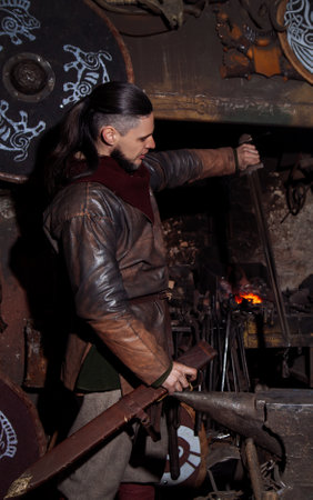 Viking forges weapons and swords in the smithy. A man in a warrior's clothes is in the smithy on the background of fire.の写真素材