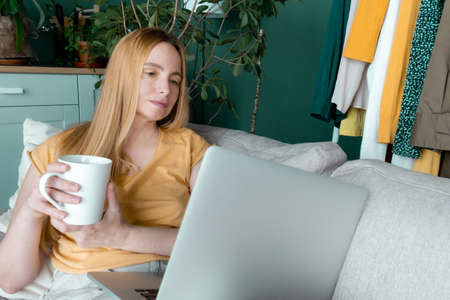 Middle Aged Blond Woman sitting on the sofa and communicates online by Laptop.の写真素材