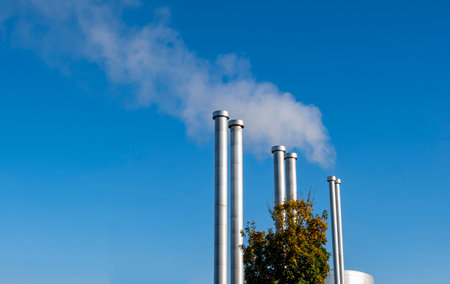 Several large metal pipes produce white steam against the background of the blue sky. Pipes of the heating boiler that gives heat to the houses.の写真素材