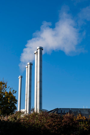 Several large metal pipes produce white steam against the background of the blue sky. Pipes of the heating boiler that gives heat to the houses.の写真素材