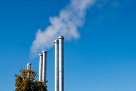 Several large metal pipes produce white steam against the background of the blue sky. Pipes of the heating boiler that gives heat to the houses.の写真素材