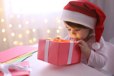 Little Christmas girl in a Santa hat looking into a gift box. Curious child with a New Years gift. The background is defocused light. Concept of Christmas and New Year 2022. High quality photoの写真素材