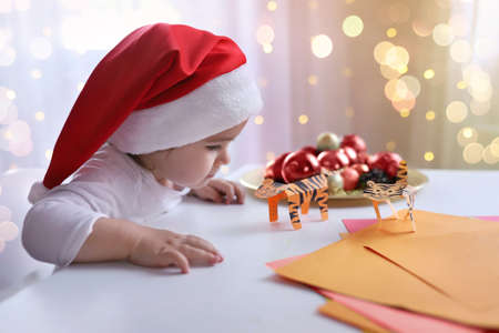 little Christmas girl sitting in New Year hat at a table with paper tigers with symbol of the year. Childrens DIY concept. Background defocus light. Handcraft creative idea for xmas new year 2022.の写真素材