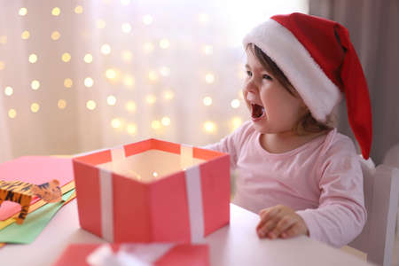 Emotional little Christmas girl in a Santa hat looks into a gift box. Child is happy with a New...
