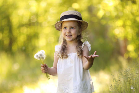 summer portrait of a little beautiful curly girl in a straw hat blowing dandelions. allergy free concept.の写真素材