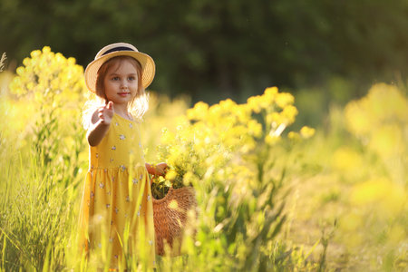 Little girl smiling on summer field in straw hat and holding bag with yellow wildflowers in her hands. Child is having fun on the street. Concept of healthy child without allergies. High quality photoの写真素材