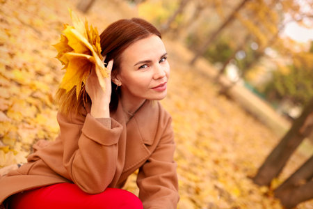 Beautiful elegant woman in autumn park holds an autumn yellow leaves near the face. Young woman in a beige coat walks outdoors in the park, concept autumn. High quality photoの写真素材
