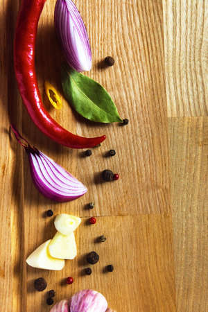 Whole chili peppers, bay leaf, yellow hot pepper slice, purple onion slice, minced garlic clove, peppercorn mixture lie on a wooden cutting board.の写真素材