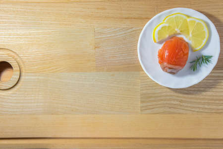 Roll of red fish with lemon wedges and juniper sprigs on a round plate on a wooden backgroundの写真素材