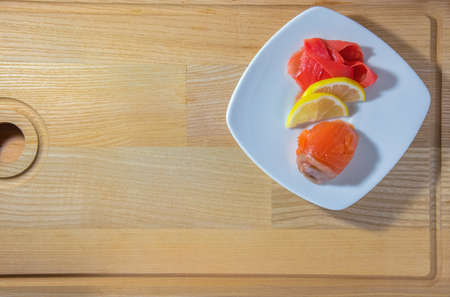 Rolled up red fish roll with ginger and lemon wedges on a white square plate and wooden backgroundの写真素材