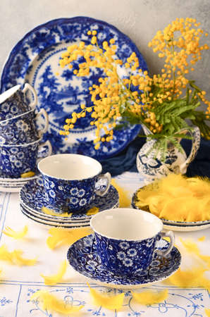 Cups with a blue pattern on the table, yellow flowers, mimosa in a vase, antique porcelain, white tablecloth, selective focus.の写真素材