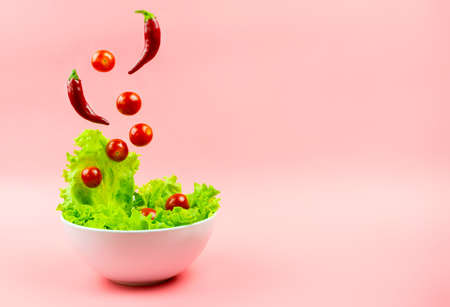 Salad, cherry tomatoes and chili peppers flying out of the white bowl on a pink background. Concept of a healthy diet. Selective focus, copyspace.の写真素材
