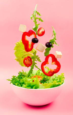 Salad ingredients falling into the bowl, pink background. Cut pepper, cherry tomatoes, olives, tofu cheese and arugula falling in the bowl. Concept of a healthy diet or cooking. Selective focus.の写真素材